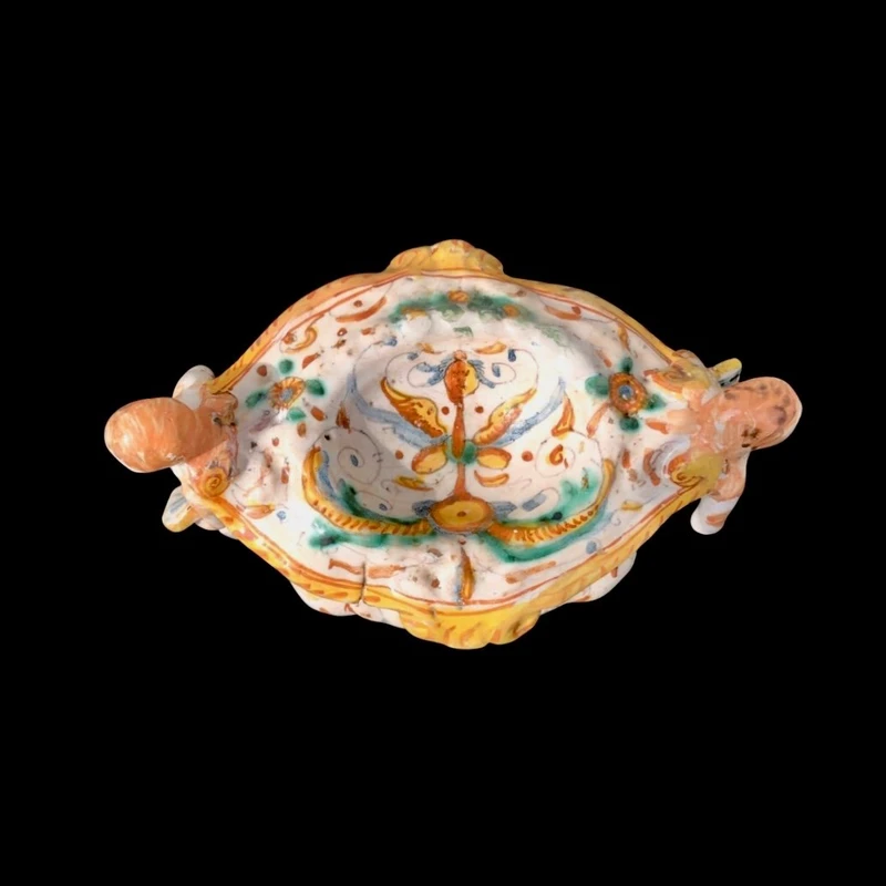 Saliera in maiolica con prese a forma di angeli che reggono dei libri e decoro a ‘raffaellesche
