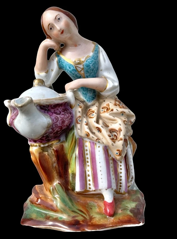 Decorated porcelain teapot-shaped nightlight with a lid shaped like a seated lady holding a teapot.