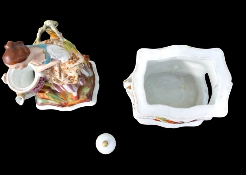 Decorated porcelain teapot-shaped nightlight with a lid shaped like a seated lady holding a teapot.