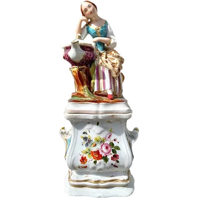 Decorated porcelain teapot-shaped nightlight with a lid shaped like a seated lady holding a teapot.