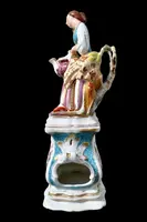 Decorated porcelain teapot-shaped nightlight with a lid shaped like a seated lady holding a teapot.