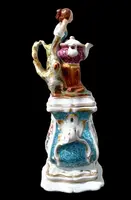 Decorated porcelain teapot-shaped nightlight with a lid shaped like a seated lady holding a teapot.
