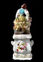 Decorated porcelain teapot-shaped nightlight with a lid shaped like a seated lady holding a teapot.
