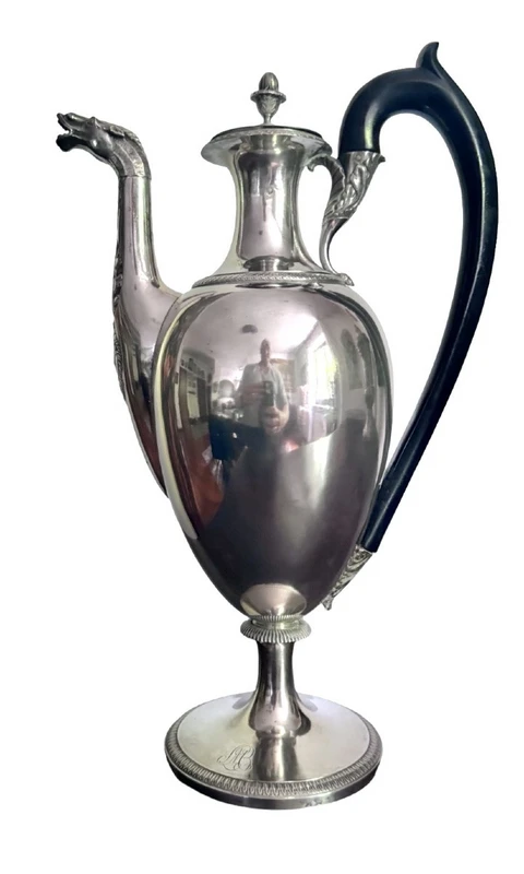 A silver coffee pot with a pear-shaped body on a circular base adorned with lanceolate leaves.